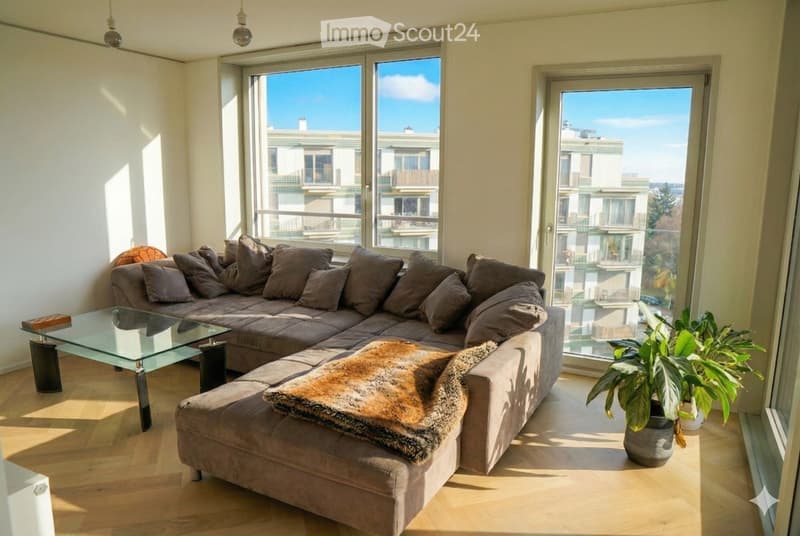 Bright living room with large windows and a balcony door offering a view of the city. Features a grey sectional sofa with multiple cushions, a glass coffee table, a fur throw, and a potted plant near the window.