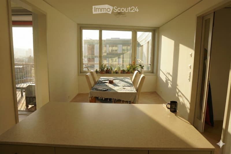 Bright dining area with a table, chairs, and potted plants on the windowsill. A coffee mug sits on a counter, and a balcony is visible through the open door.
