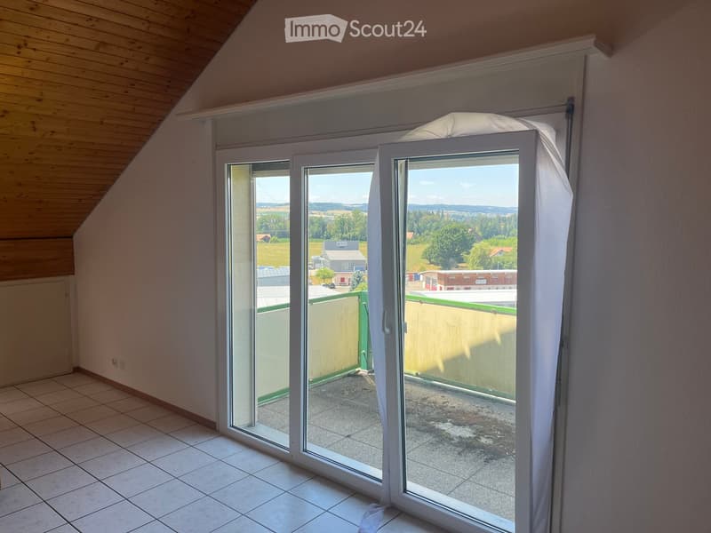 Empty room with tiled floor, wooden slanted ceiling, large sliding glass doors leading to a balcony with a view of the countryside.