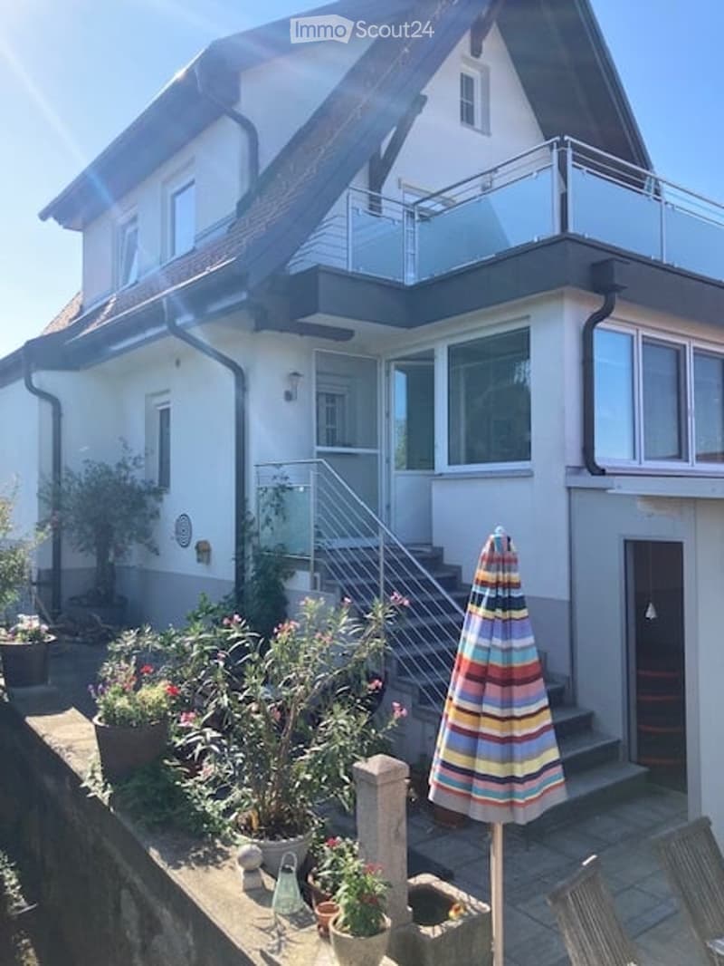 Two-story house with a balcony, large windows, and a well-maintained garden area with potted plants and an umbrella.