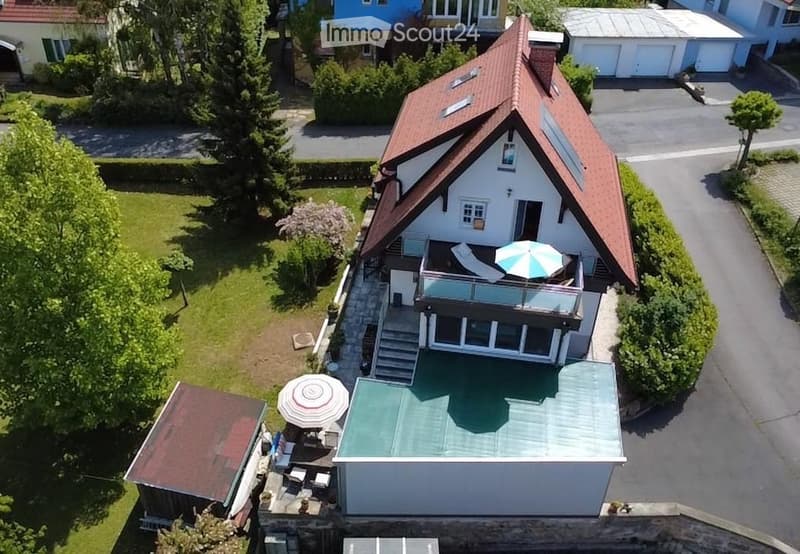 Aerial view of a two-story house with a red-tiled roof, white exterior, and a balcony with an umbrella. The property includes a well-maintained lawn, a patio area, and neighboring houses in the background.
