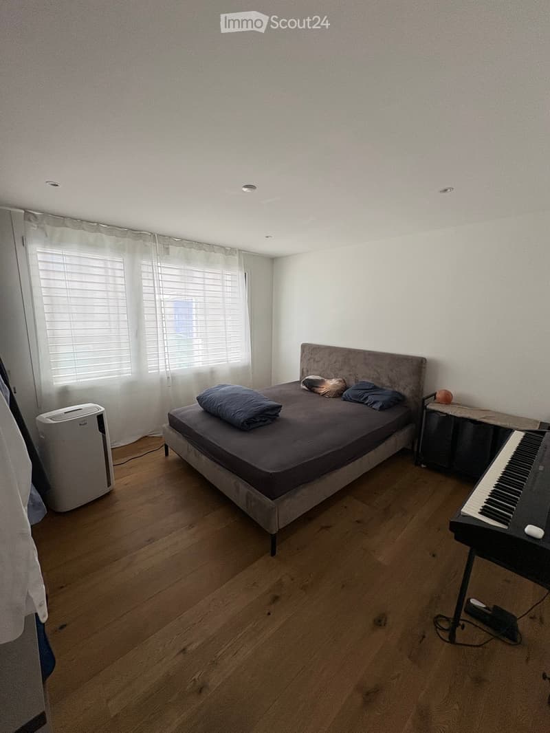 Bedroom with wooden flooring, a bed with pillows, a piano, an air conditioner, and a small table with a fruit bowl.