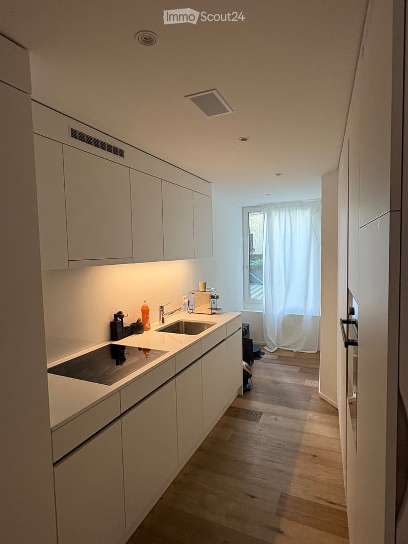 Modern kitchen with white cabinets, wooden flooring, and a small window with white curtains. Features a sink, faucet, coffee maker, and induction cooktop.