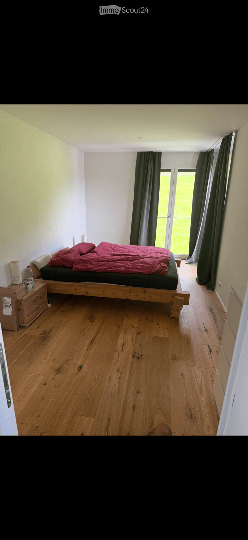 Minimalist bedroom with wooden flooring, a low platform bed with red bedding, a small nightstand, and large windows with dark curtains.