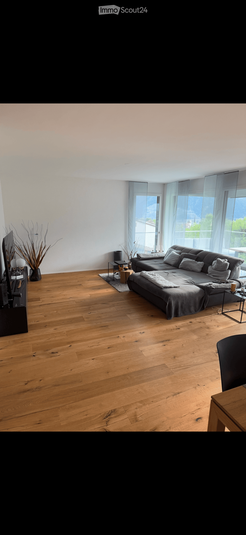Modern living room with wooden flooring, large windows, grey sectional sofa, coffee table, side tables, and a television stand.