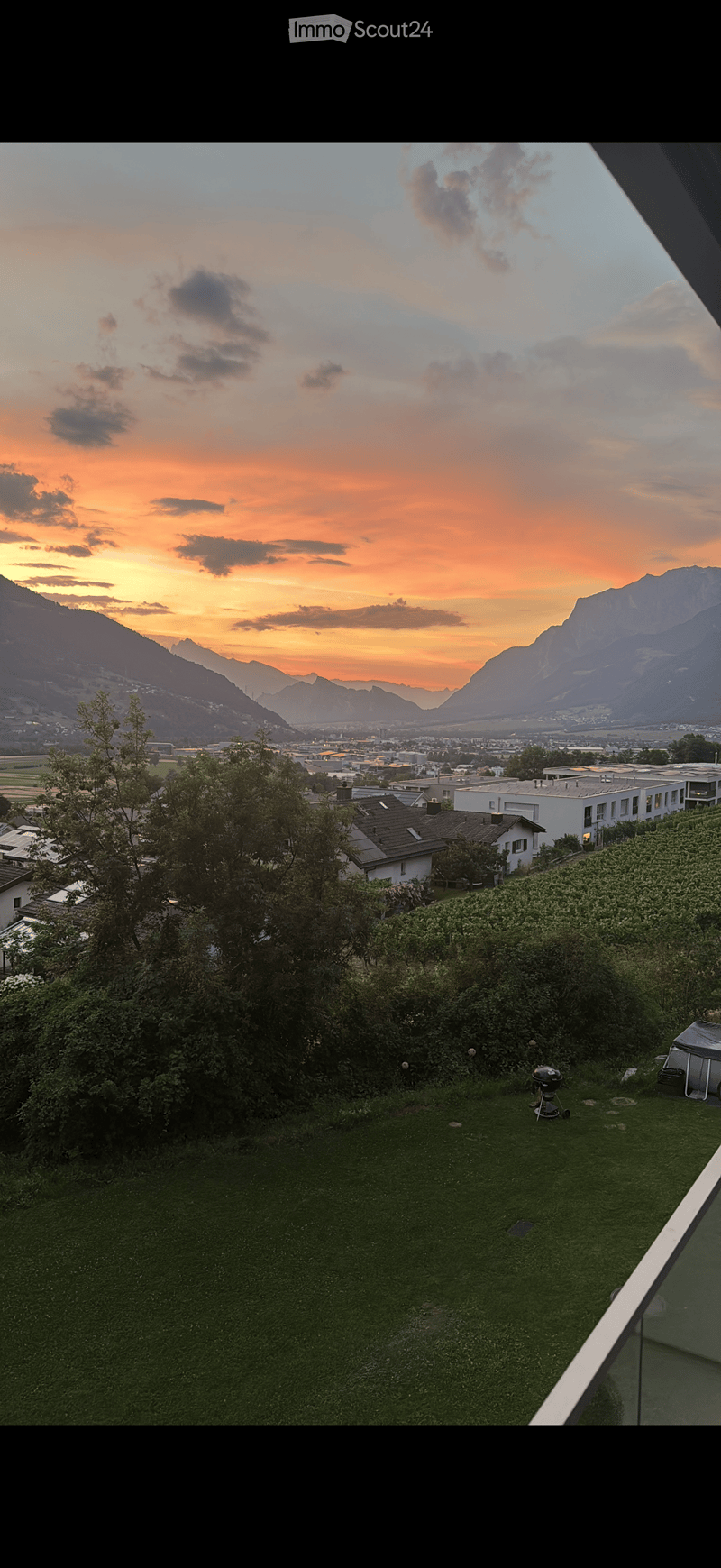Garden with green grass, trees, and a view of mountains and cityscape at sunset.