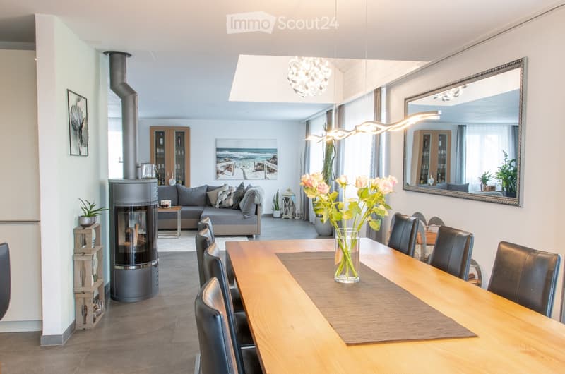 Open-plan living and dining area with a grey sectional sofa, wooden dining table with black chairs, modern fireplace, large mirror, and decorative vases with flowers.