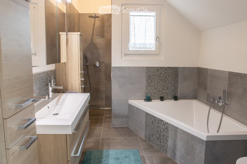 Modern bathroom with a white sink, bathtub, shower, and wooden cabinet. Features a large mirror, decorative tiles, and a small window with a curtain.