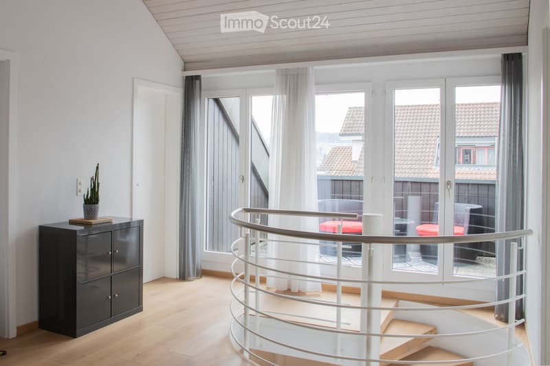 Spacious, modern room with light wooden flooring, white walls, and large windows with sheer curtains. A sleek black cabinet with a potted plant sits near the wall, and a curved white staircase with a glass railing leads upwards.