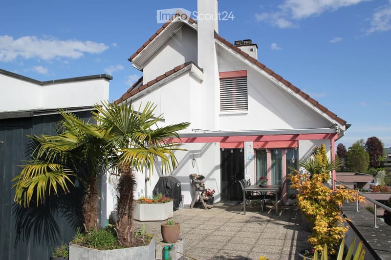 A modern white house with a red-tiled roof and a small balcony area. The balcony features a table, chairs, potted plants, and a palm tree. The house has a chimney and a covered entrance with hanging lights.