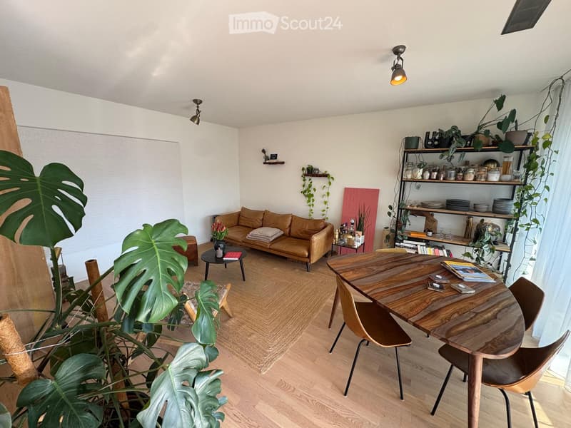 A modern living room with a brown leather sofa, wooden coffee table, dining table with chairs, shelves with plants and decorative items, and large leafy plants in the corner.