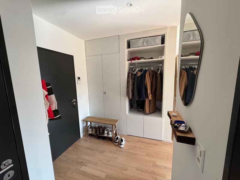Modern hallway with light wooden flooring, built-in white closet with hanging clothes, a small wooden bench holding shoes, and a wall-mounted shelf with items. A large oval mirror is mounted on the wall.