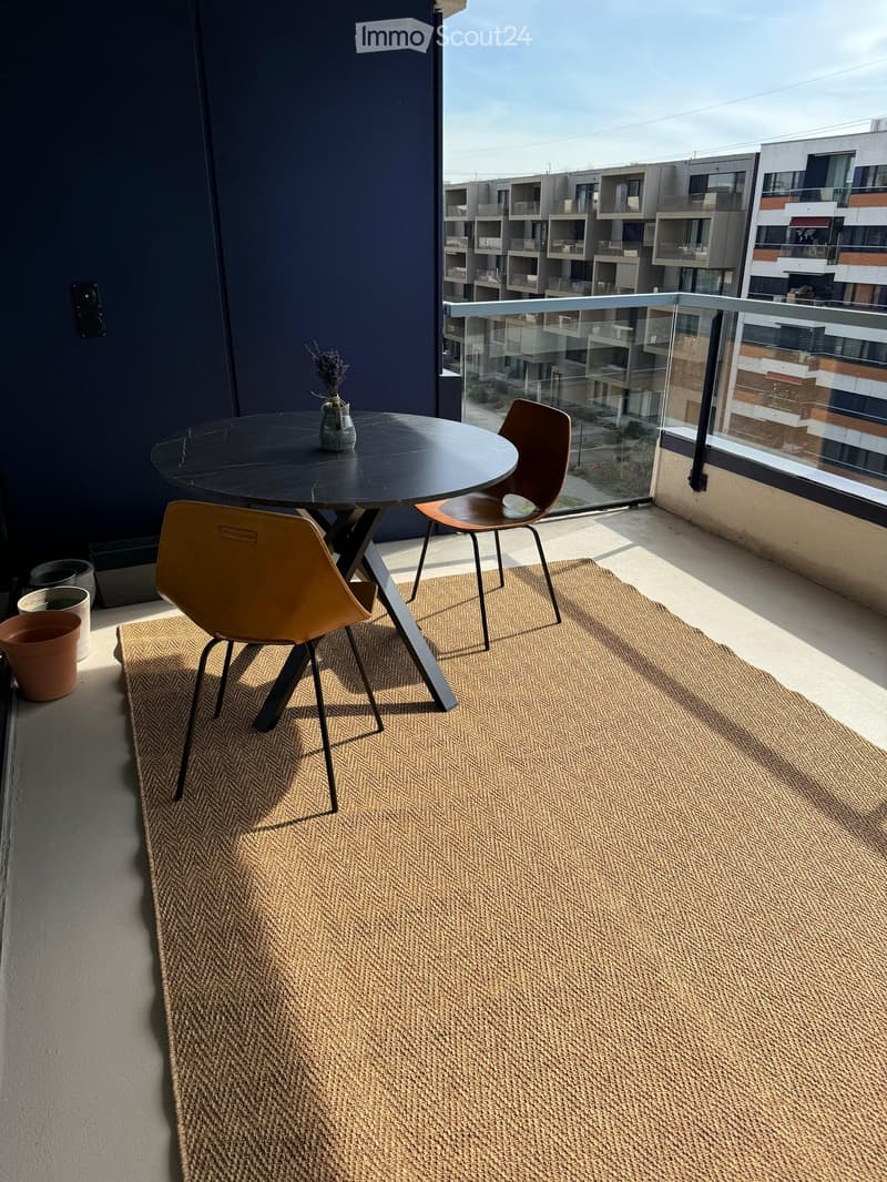 Balcony with a round black table, two modern chairs, a small vase with dried flowers, a large woven rug, and a glass railing overlooking a modern apartment complex.
