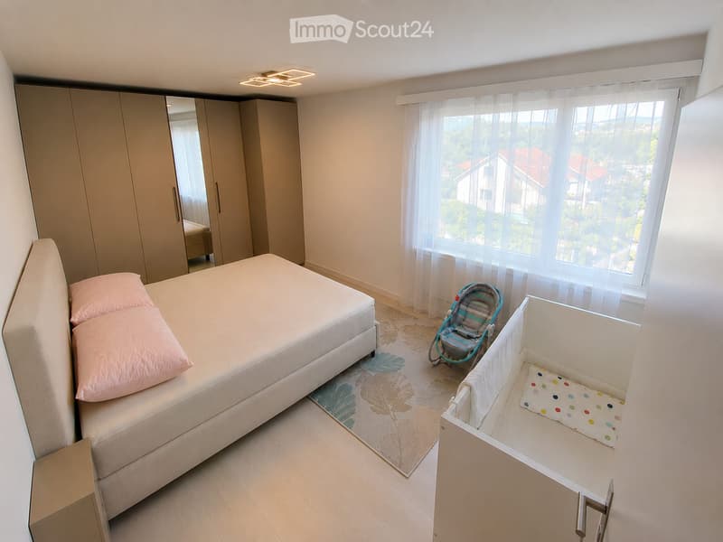 Bright bedroom with a large bed, pink pillows, a baby stroller, a crib, and a modern wardrobe. Large windows offer a scenic view.