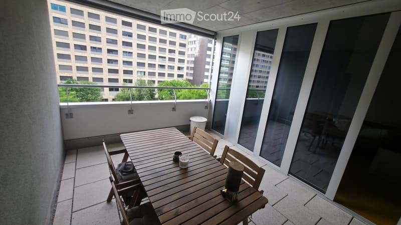 Balcony with wooden table, chairs, and a view of a cityscape and greenery.
