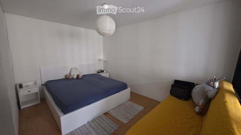 A small, modern bedroom with a white bed, blue bedding, a nightstand, and a hanging paper lamp. The room features wooden flooring and a colorful rug.