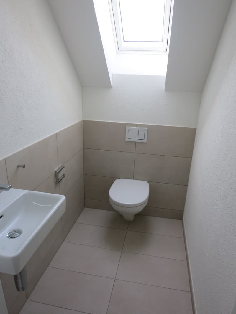 Modern bathroom with a toilet, sink, tiled walls, and a small window at the top.
