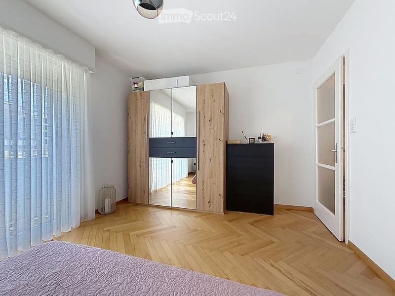 Modern bedroom with wooden flooring, a large mirrored wardrobe, a nightstand with a lamp, and a neatly made bed.