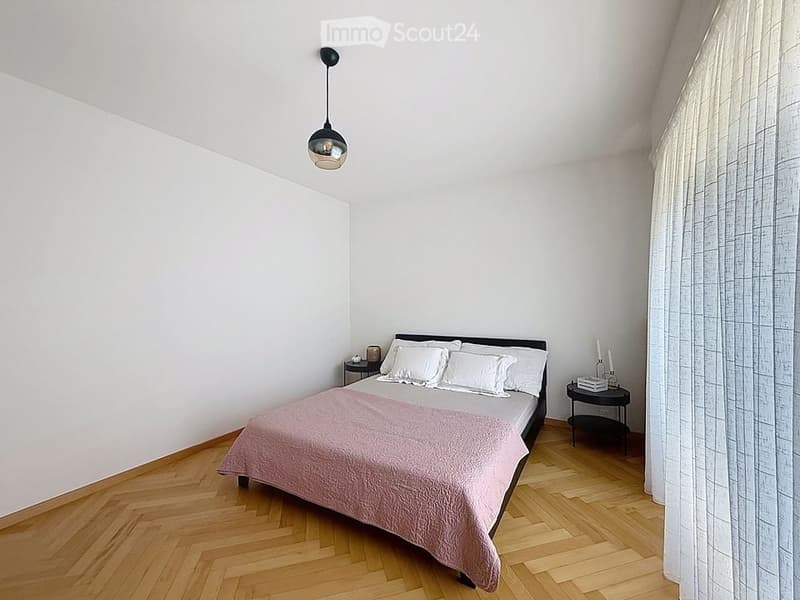 Minimalist bedroom with a wooden floor, white walls, a single bed with white pillows and a pink blanket, a nightstand with a lamp, and a curtain on the window.