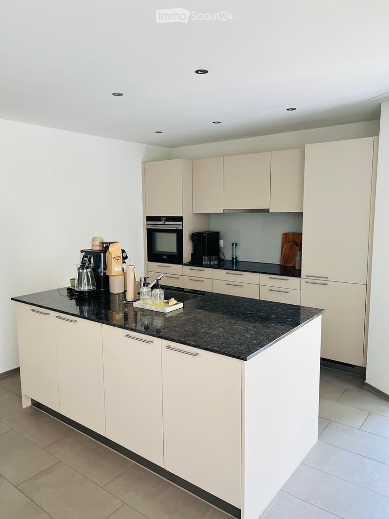 Modern kitchen with light-colored cabinets, granite countertop, and various appliances including a coffee maker, oven, and cutting board.