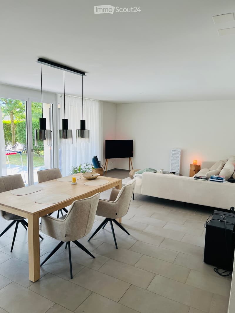 Modern living and dining area with a white sofa, wooden dining table, chairs, and a television. The space features large windows with white curtains and pendant lights.
