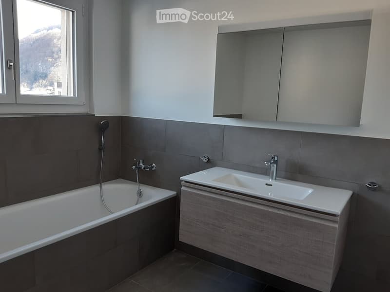 Modern bathroom with a bathtub, sink, mirror, and grey tiled walls.