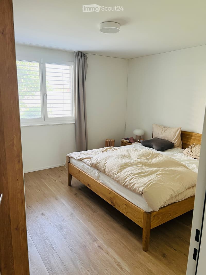 A modern bedroom with a wooden bed, beige bedding, a nightstand with a lamp, and a window with blinds and curtains.