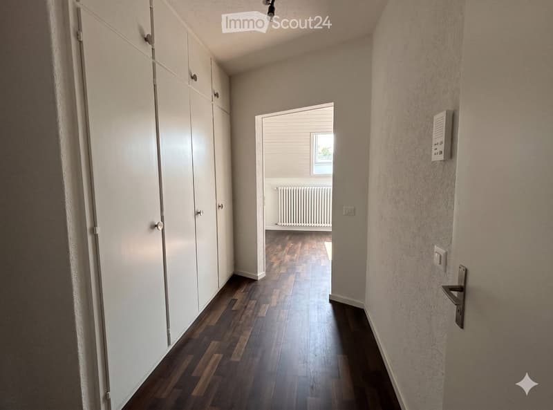 A narrow hallway with wooden flooring, white walls, and a closed white cabinet on the left. A door at the end leads to another room with a radiator and a window.