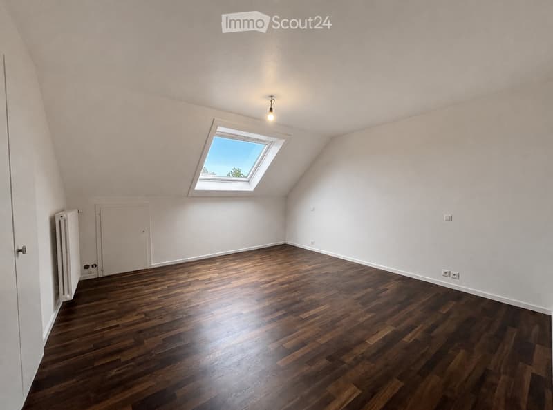 Empty attic room with wooden flooring, white walls, a skylight window, and a radiator. No furniture or equipment present.
