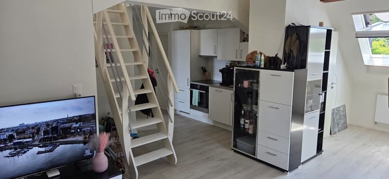 Modern, open-plan living and kitchen area with a spiral staircase leading to an upper level. Features white kitchen cabinets, stainless steel appliances, a large TV, and a decorative vase on a table.