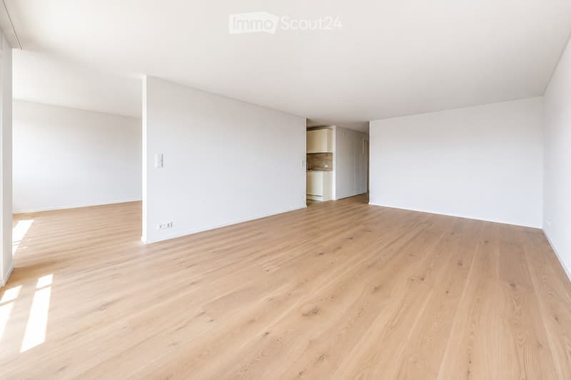 Empty room with light wooden flooring and white walls, featuring a small adjacent kitchen area with cabinets and appliances.