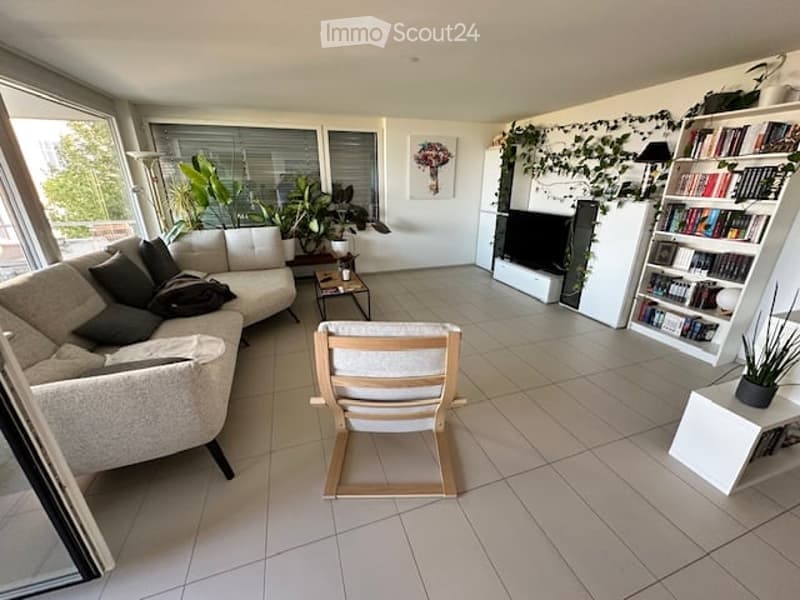 Bright living room with large windows, white tiled floor, grey sectional sofa, wooden coffee table, armchair, bookshelf, TV, and indoor plants.