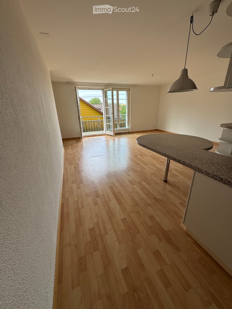 Empty room with wooden flooring, white walls, and a large open doorway leading to a balcony or outdoor area. A small round table is visible near the doorway.