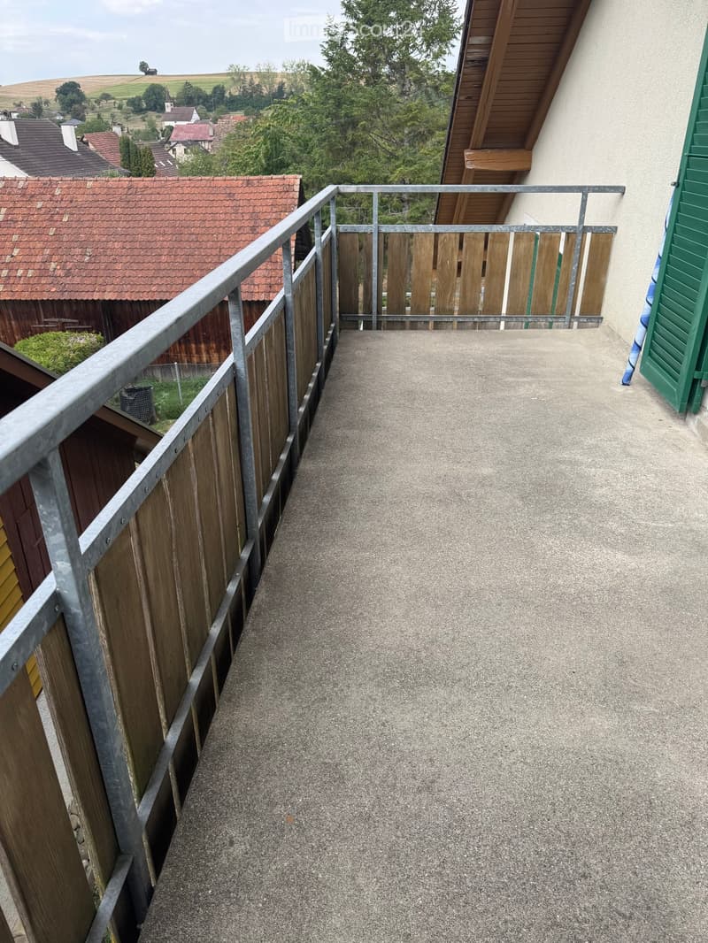 Balcony with metal railing and wooden fence, overlooking a rural landscape with houses and greenery.