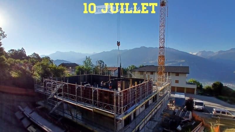 A multi-story building under construction with scaffolding, surrounded by trees and mountains, with a crane lifting materials.