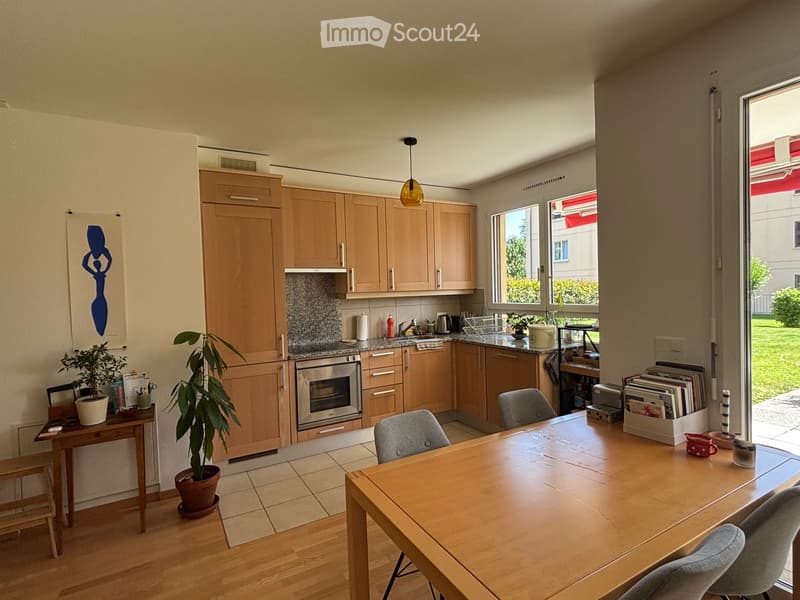 The image shows a modern kitchen and dining area with light wooden cabinets, a central island, and a dining table with chairs. There are potted plants, a sink, and various kitchen appliances visible.