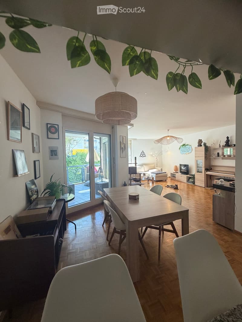 Open-plan living and dining area with wooden flooring, large windows, and modern furniture including a dining table, chairs, sofa, TV, and piano.