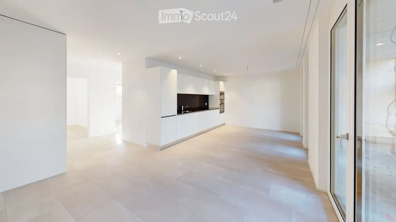 Bright, open-plan living and dining area with a modern white kitchen featuring cabinets, a stove, and a sink. The space has large windows and a glass door leading to a balcony.