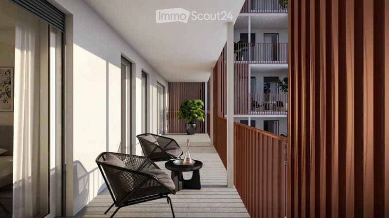 Modern balcony with two wicker chairs, a small round table, and a potted plant. The balcony has wooden railings and is part of a multi-story building with additional balconies visible in the background.