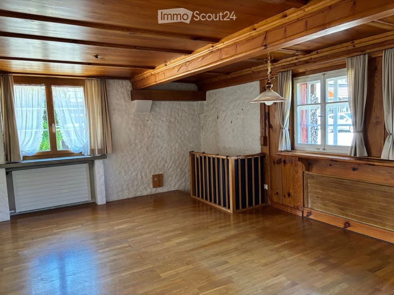 Empty room with wooden floors, exposed wooden beams on the ceiling, large windows with curtains, and a radiator under one window.