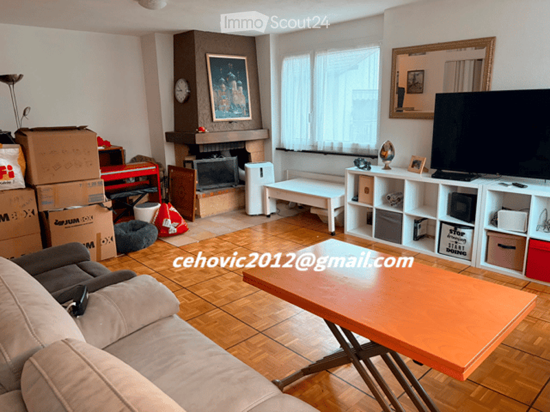 Living room with wooden flooring, fireplace, large window with white curtains, sofa, coffee table, TV stand, shelf, and a small white bench.