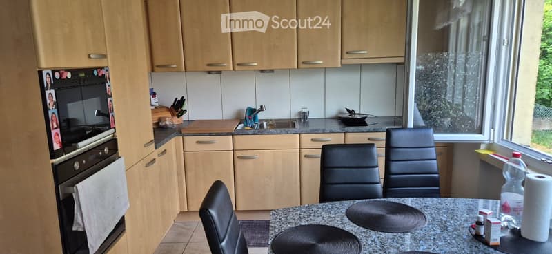 Modern kitchen with light wood cabinets, stainless steel appliances, granite countertop, and four black chairs around a dining table.