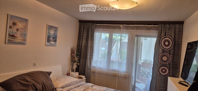 Cozy bedroom with a bed, nightstand, and large window leading to a balcony. Decor includes wall art, a curtain, and a decorative wall hanging.
