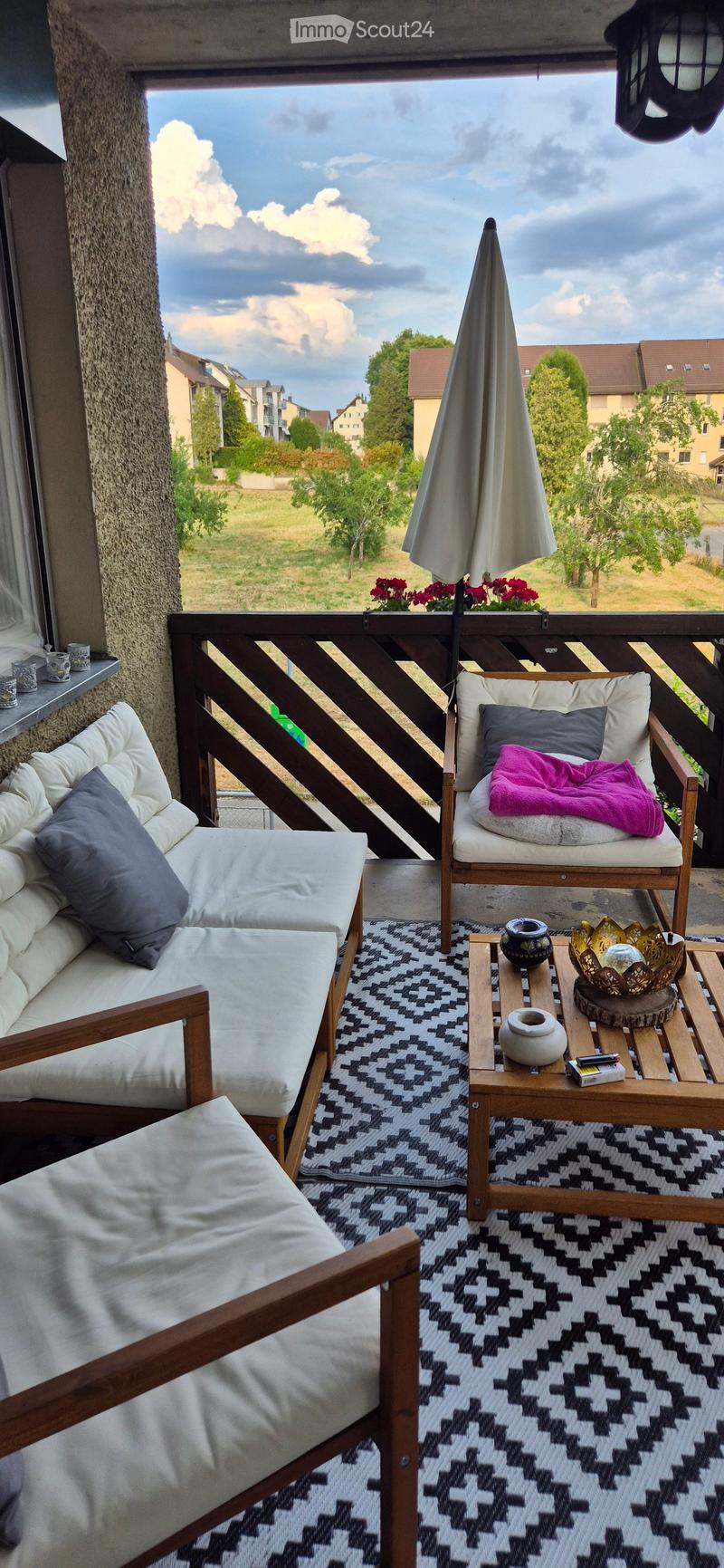 Balcony with a sofa, chair, small table, umbrella, and patterned rug, overlooking a residential area with trees and houses.