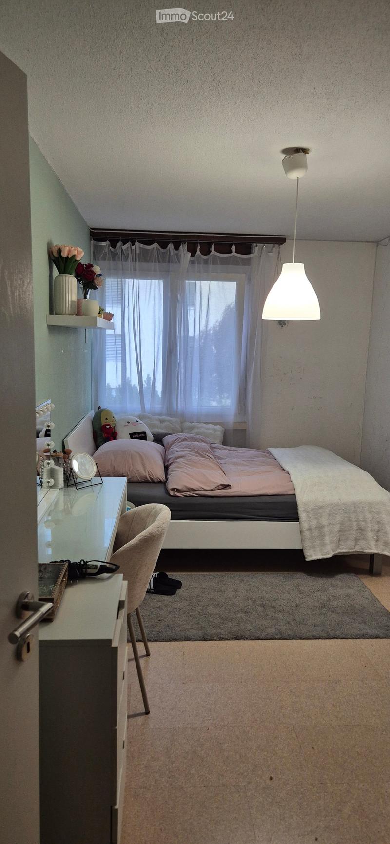Cozy bedroom with a bed, pink bedding, a chair, a mirror, a small table with a vase, and a hanging lamp.