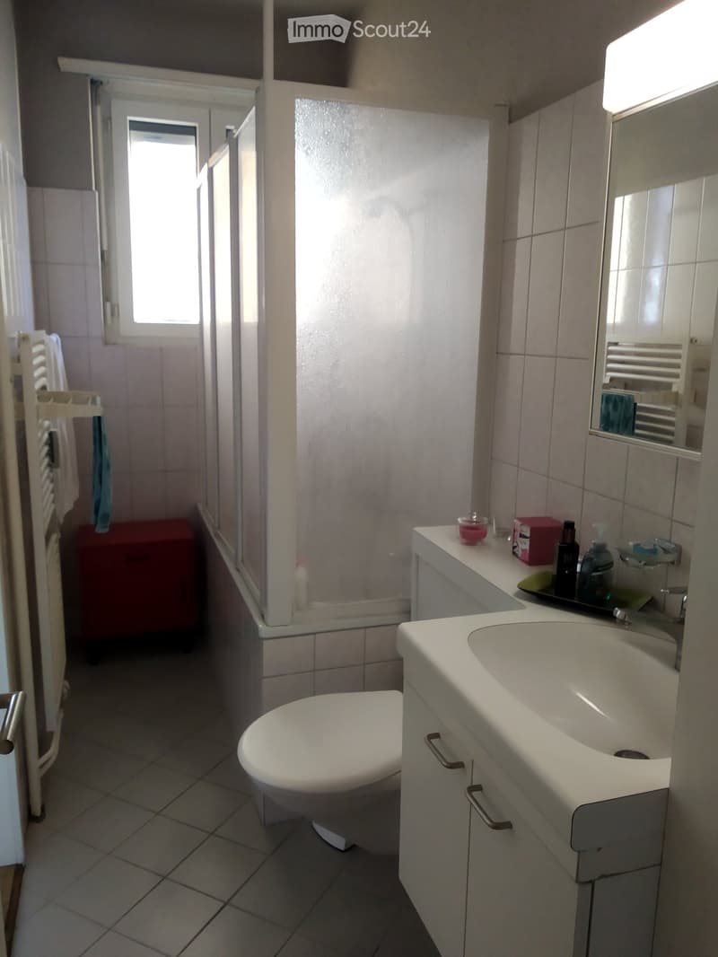 A small bathroom with a white sink, toilet, mirror, and cabinet. The sink has a faucet and a small cabinet below. A towel rack with a towel is visible on the left side, and a window provides natural light.