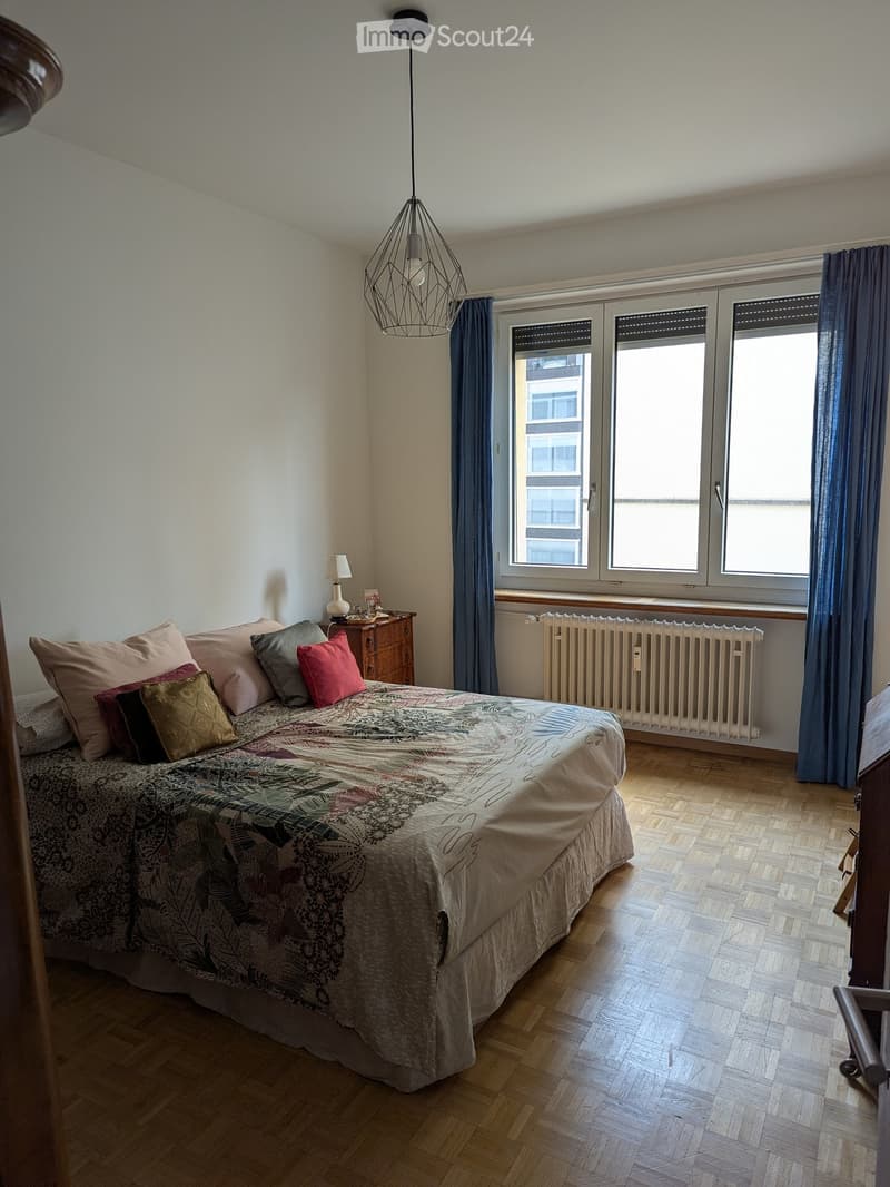 Cozy bedroom with wooden flooring, large window with blue curtains, bed with patterned duvet and multiple pillows, nightstand with lamp, radiator below window, and modern geometric pendant light.