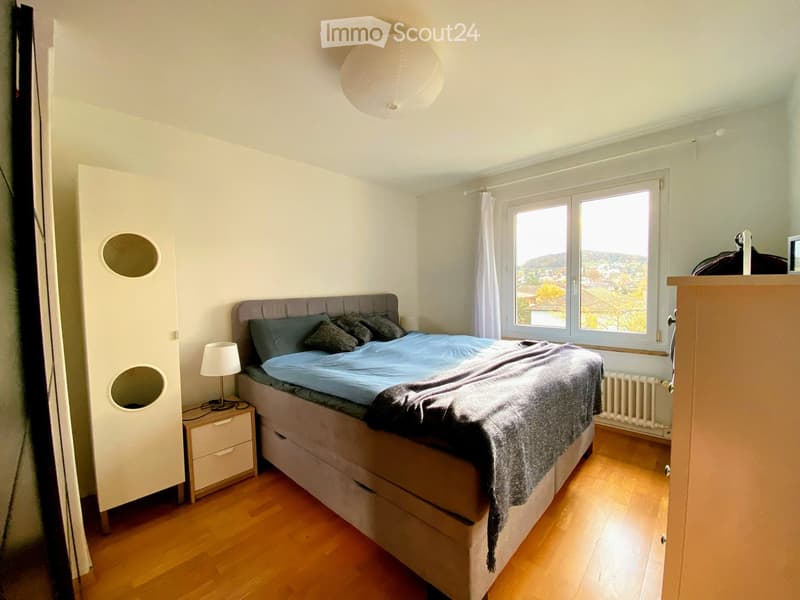 Cozy bedroom with wooden flooring, large window offering a scenic view, a neatly made bed with blue bedding, a nightstand with a lamp, and a modern wardrobe.