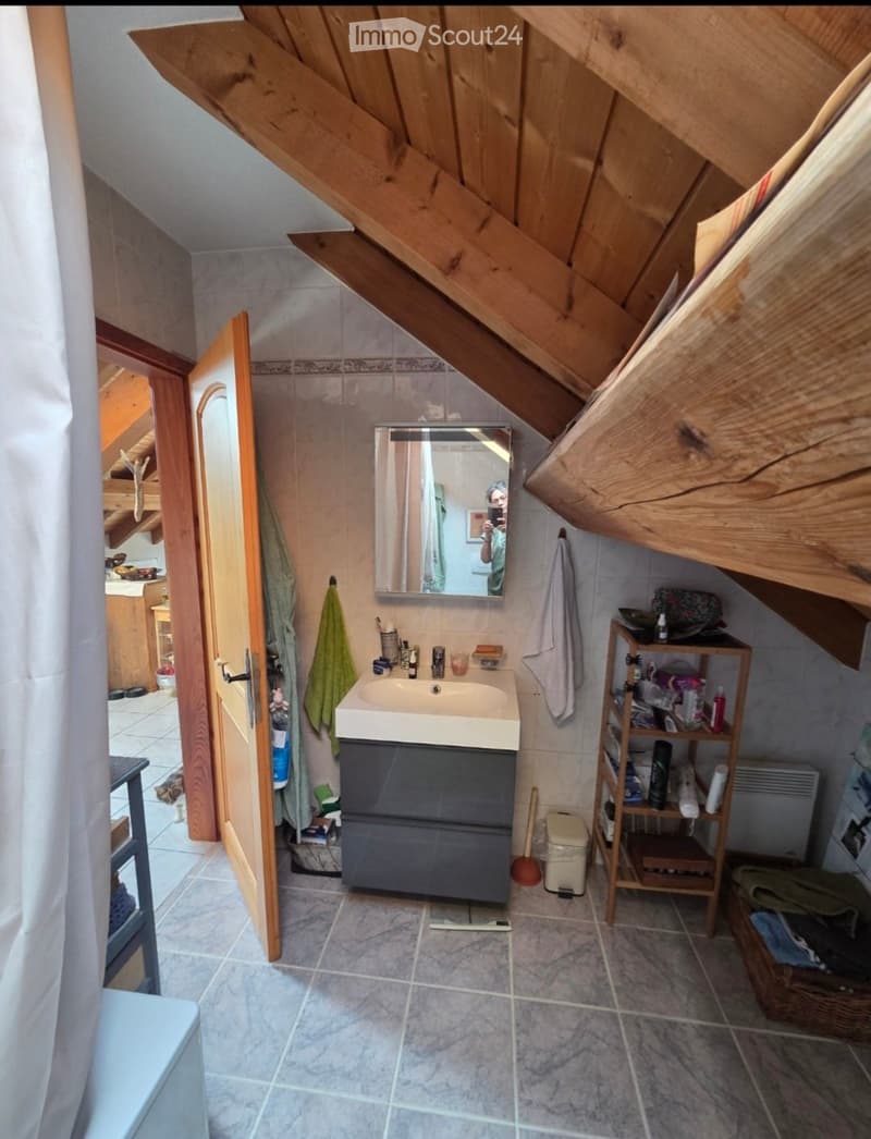 Bathroom with a sink, mirror, cabinet, shelf, towels, toilet, and radiator. The room has exposed wooden beams on the ceiling and tiled flooring.