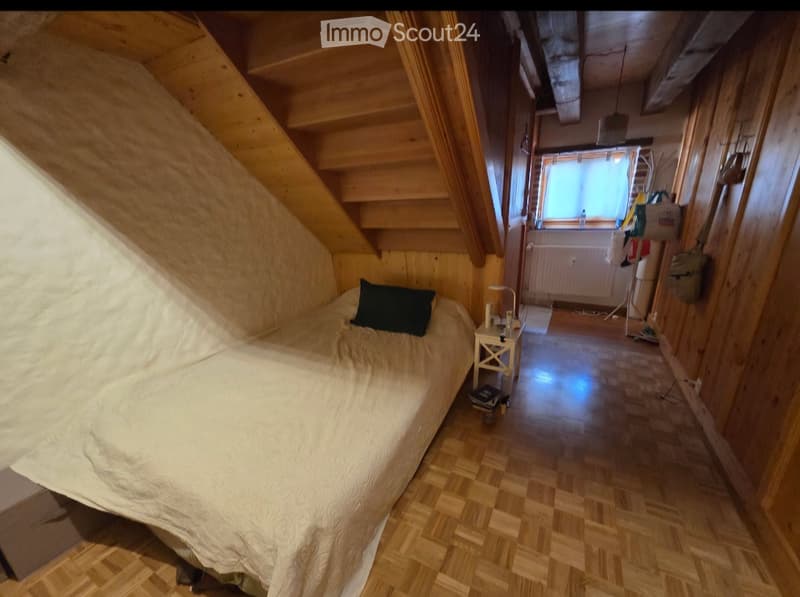 Cozy attic bedroom with wooden walls and slanted ceilings, featuring a single bed, nightstand, and a view of a hallway leading to another room.